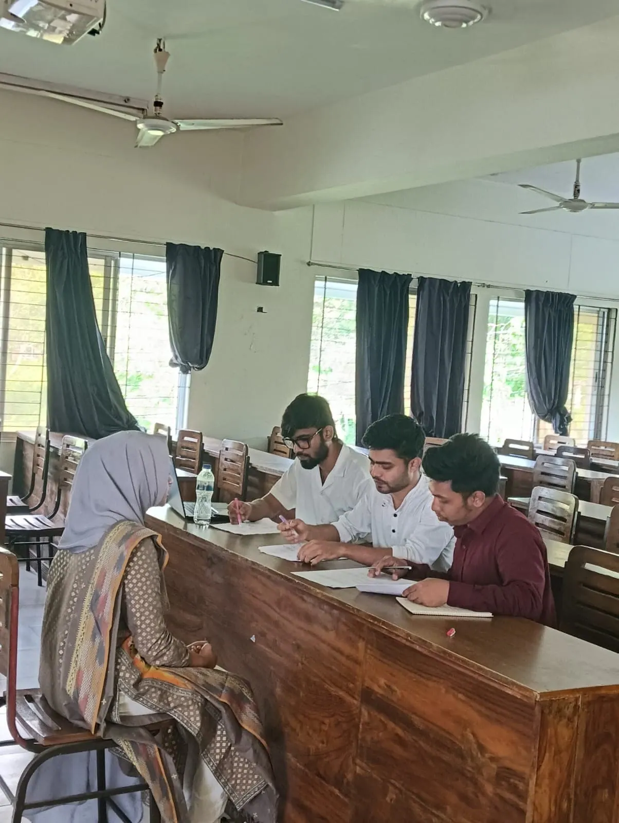 BioPC mentors working through a problem with a student across a classroom desk at the University of Chittagong